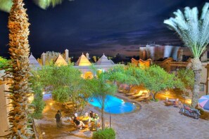 A pool area with lounge chairs and umbrellas, surrounded by palm trees and a building with multiple domes. - Riad Jnane Ines (Machraa El Ain)