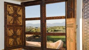 A room with a checkered floor, a large window with a view of a landscape, and a wooden cabinet with intricate designs. - Riad Jnane Ines (Machraa El Ain)