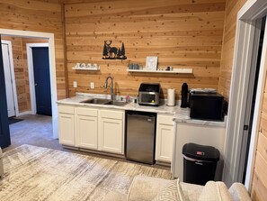 A compact kitchen with a sink, microwave, and refrigerator, wooden walls, and a small trash bin.