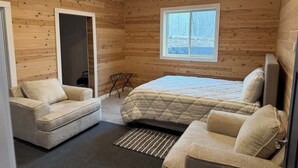Um quarto de cabana de tronco com uma cama, duas poltronas, uma janela com vista para a neve e uma porta que dá para outro cômodo.