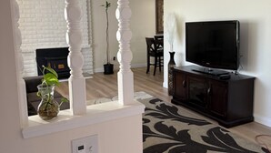 Uma sala de estar com lareira, uma TV sobre um móvel, um tapete estampado e uma planta no parapeito da janela.