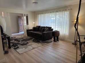 Un salón con un sofá marrón oscuro, un tapiz estampado, una lámpara de pie y una ventana con cortinas translúcidas.