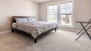 Un dormitorio con cama, dos ventanas y una mesita.