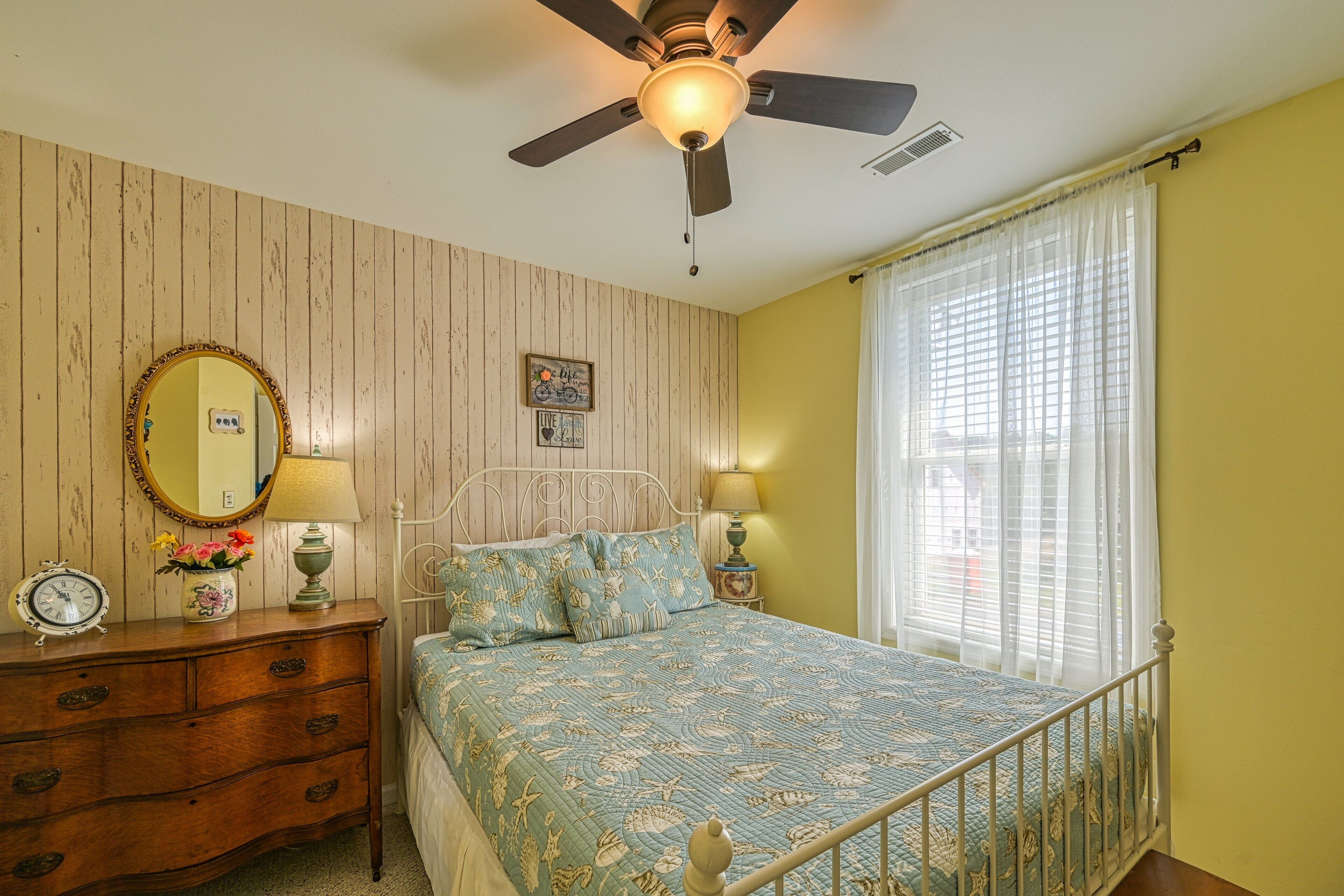 A bedroom with a bed, dresser, mirror, ceiling fan, and window with curtains.