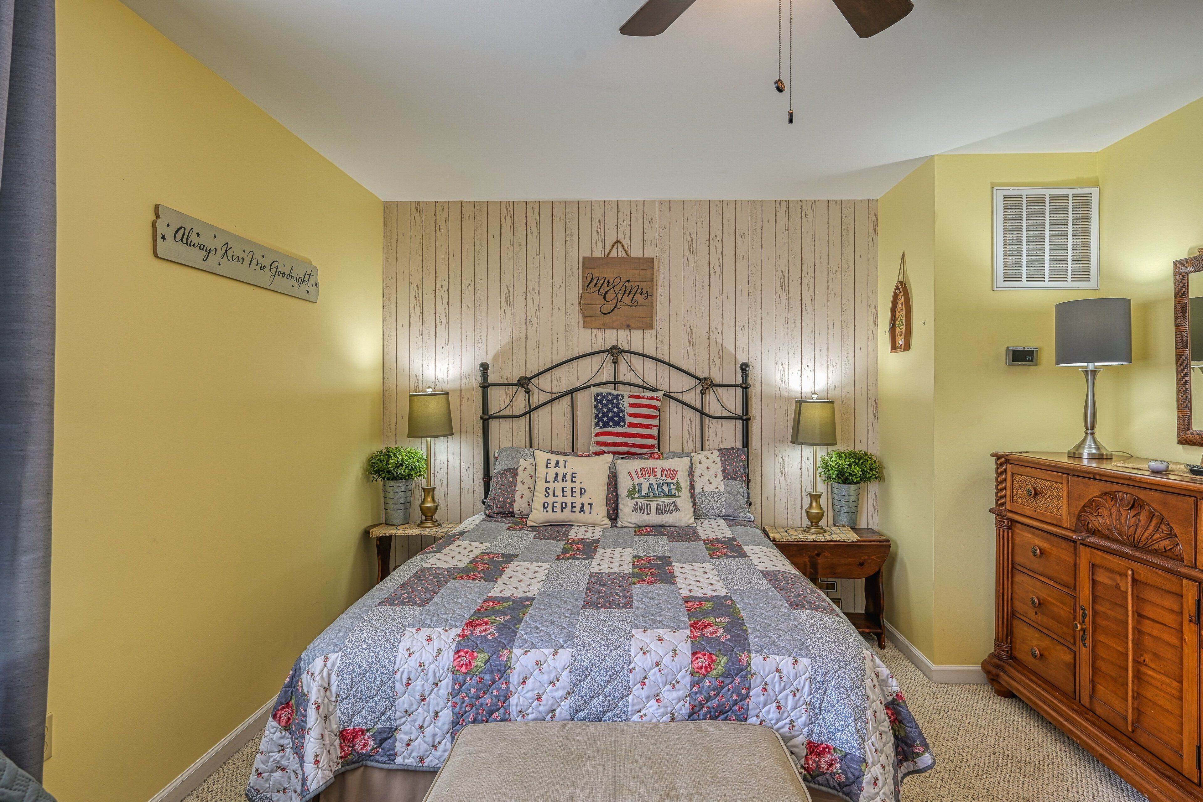 A bedroom with a bed, a nightstand, a dresser, a ceiling fan, and a wall with a sign.