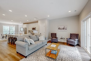 A modern living room with a sofa, coffee table, and chairs, a kitchen area with white cabinets, and a dining space with bar stools. - Charming Lakefront Retreat With Game Room and Covered Dock Access (Mineral)