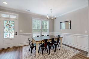 A dining area with a wooden table and chairs, a chandelier, a patterned rug, and a window with blinds. - Charming Lakefront Retreat With Game Room and Covered Dock Access (Mineral)