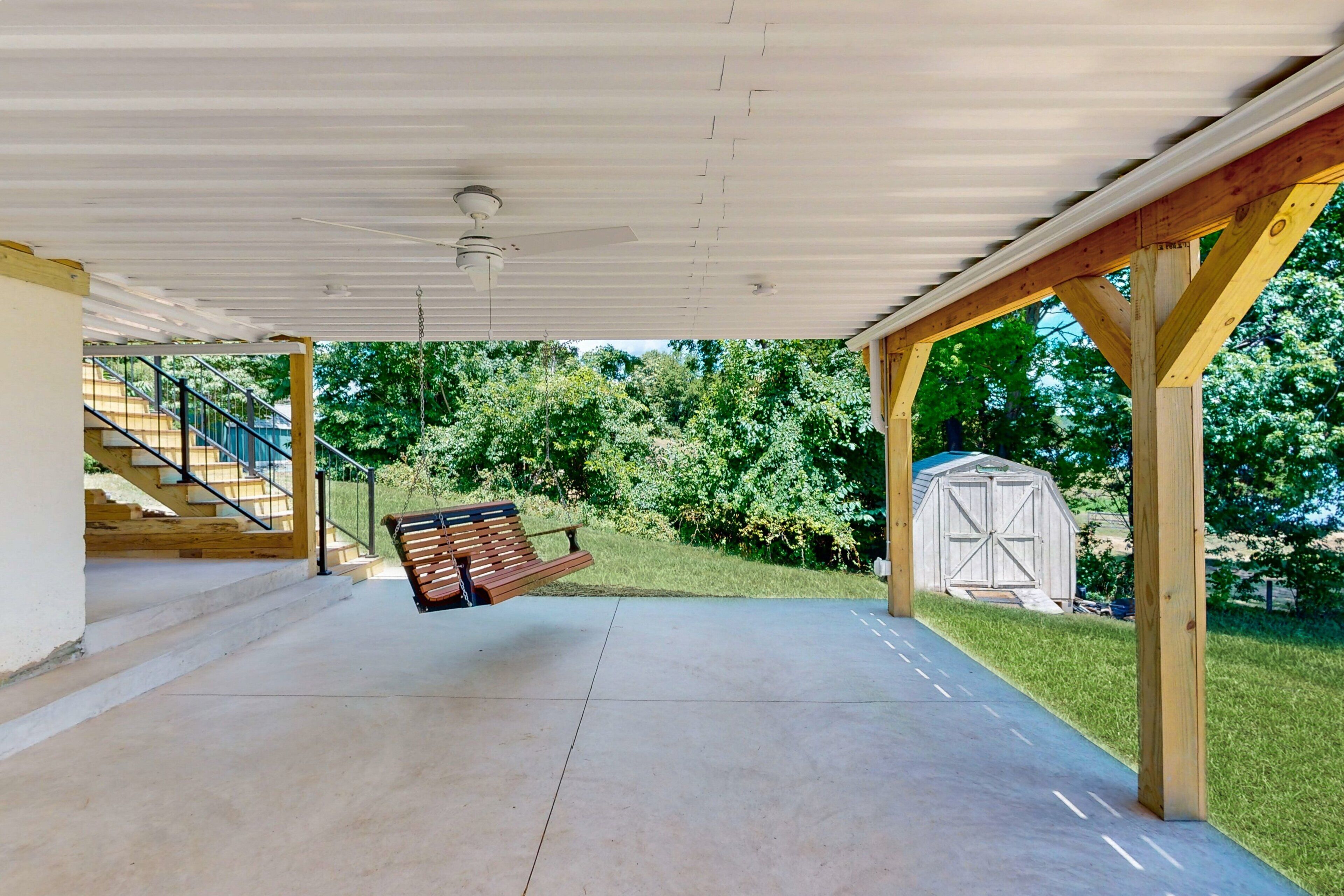 A covered patio area with a swing, a wooden bench, a concrete floor, a staircase, and a shed in the background.