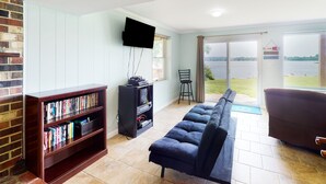 A living room with a sofa, a bookshelf, a TV, and a view of a lake through a glass door. - Charming Getaway With a Fireplace, Large Deck, Paddle Boards & Porch Swing (Mineral)