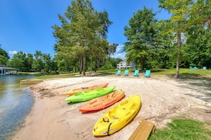 Kajaks reihen sich an einem sandigen Strand entlang, mit einem hölzernen Steg und einem Haus im Hintergrund.