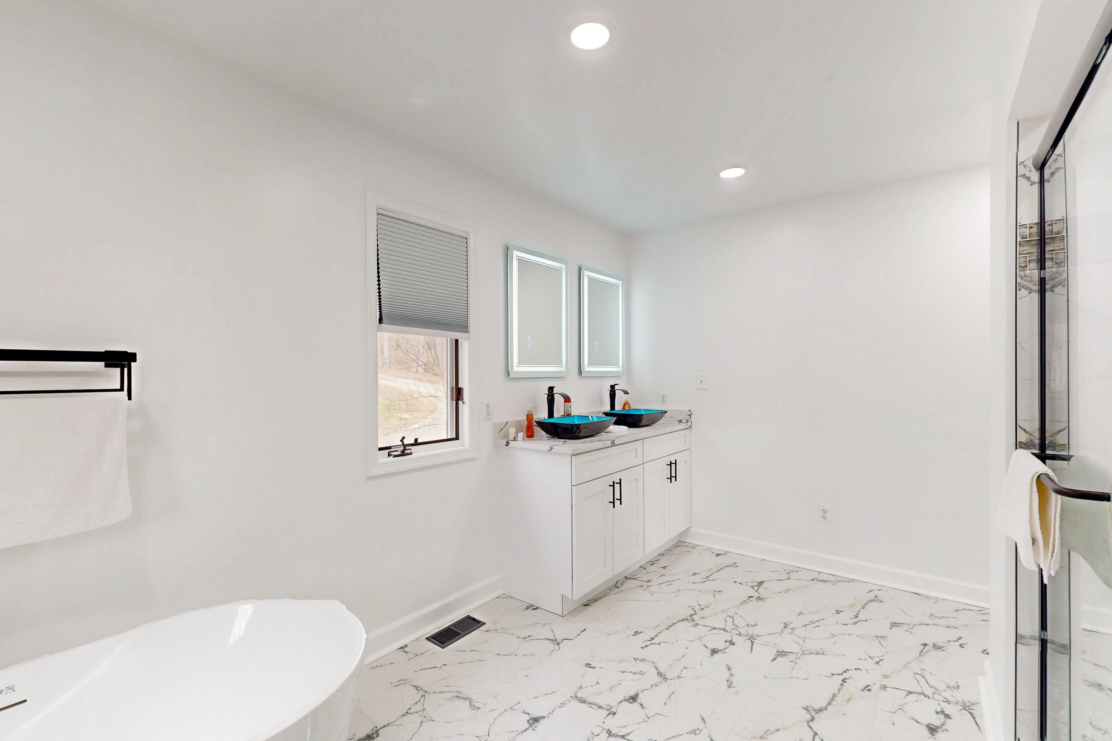 Un baño con una bañera blanca, dos lavamanos con lavabos azules, armarios blancos y una ventana con persianas.