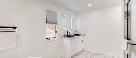 Un baño con una bañera blanca, dos lavamanos con lavabos azules, armarios blancos y una ventana con persianas.