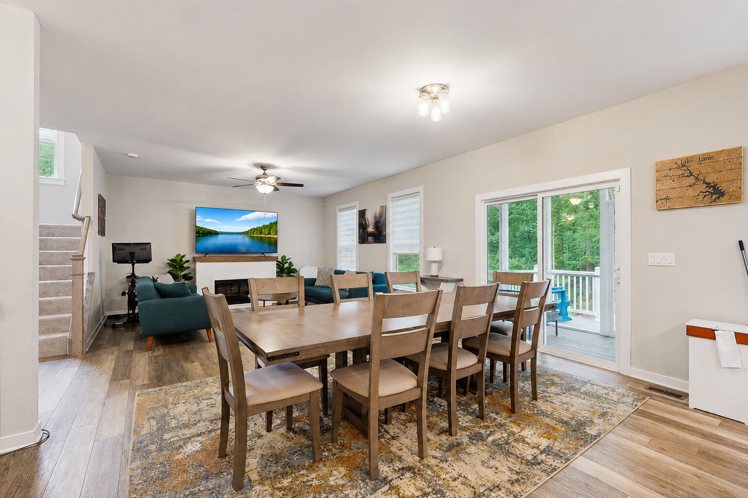 A modern living room with a dining area, a fireplace, a ceiling fan, and a large window with a view of trees.