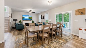 A modern living room with a dining area, a fireplace, a ceiling fan, and a large window with a view of trees. - Cozy Retreat With Forest Views, Peaceful Vibes & Modern Comfort (Bumpass)