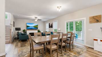 A modern living room with a dining area, a fireplace, a ceiling fan, and a large window with a view of trees.