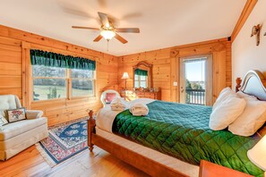 A wooden cabin bedroom with a bed, a chair, a nightstand, a lamp, and a view of the outdoors. - Dog-friendly, Lake View Cabin w/ Furnished Decks, Ping-pong & Darts (Louisa)
