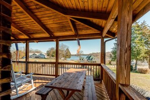 A wooden deck with a picnic table and a rocking chair, overlooking a lake and houses. - Dog-friendly, Lake View Cabin w/ Furnished Decks, Ping-pong & Darts (Louisa)