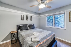 A bedroom with a bed, a ceiling fan, a window with a view of trees, and a small side table.