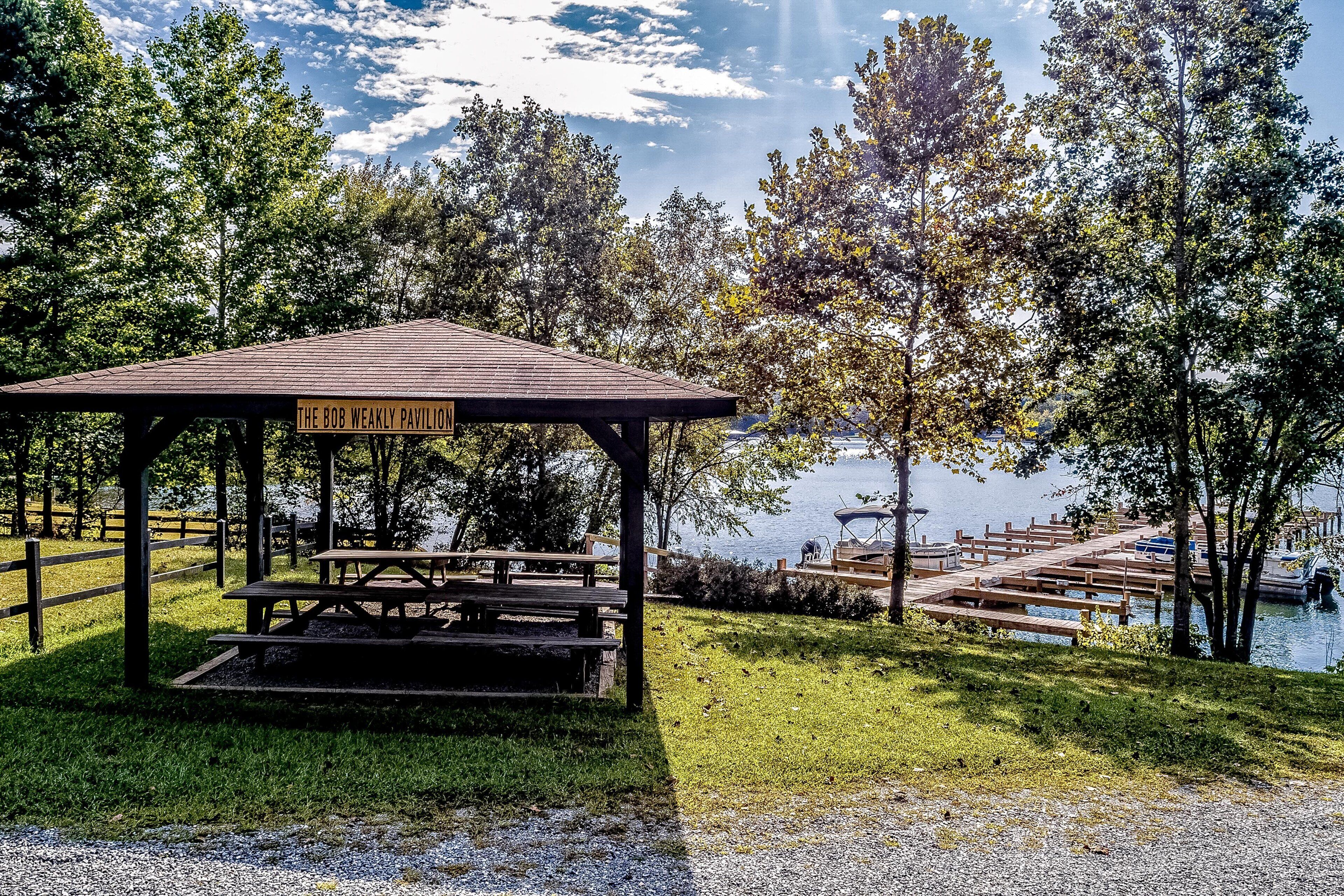 A wooden pavilion with a picnic table and a sign reading 'The Bob Weakly Pavilion' by a lake with boats and trees.