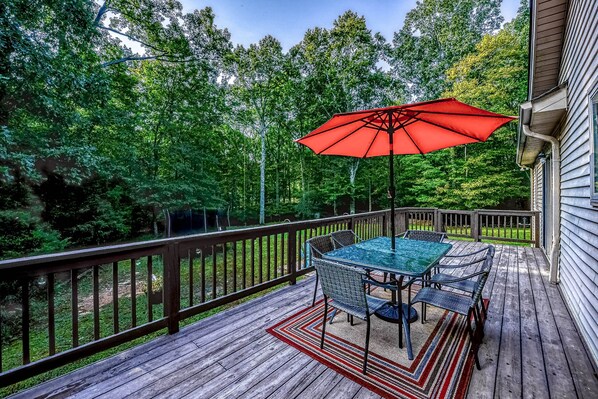 A wooden deck with a glass table and chairs, a red umbrella, and a view of a forest.