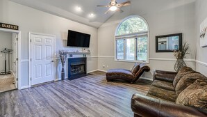 A spacious living room with hardwood flooring, a fireplace, a ceiling fan, a large window, and a television. - Dog-friendly Home With Free Wifi, Central AC, Firepit, & Furnished Deck (Mineral)