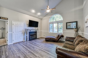 A spacious living room with hardwood flooring, a fireplace, a ceiling fan, a large window, and a television. - Dog-friendly Home With Free Wifi, Central AC, Firepit, & Furnished Deck (Mineral)