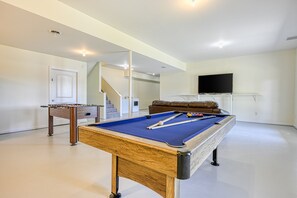 A pool table with a foosball table, a sofa, and a TV in a room with white walls and a staircase. - Dog-friendly, Lakefront Home With Private Dock, 2 Kayaks, Decks & Game Room (Mineral)