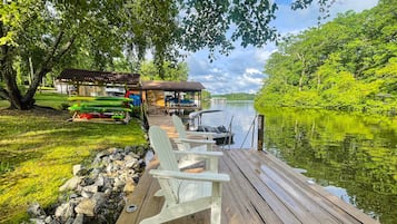 A wooden dock with Adirondack chairs, a covered seating area, and a view of a calm lake surrounded by greenery.