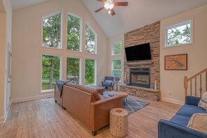 A spacious living room with large windows, a stone fireplace, a flat-screen TV, and a cozy seating area.