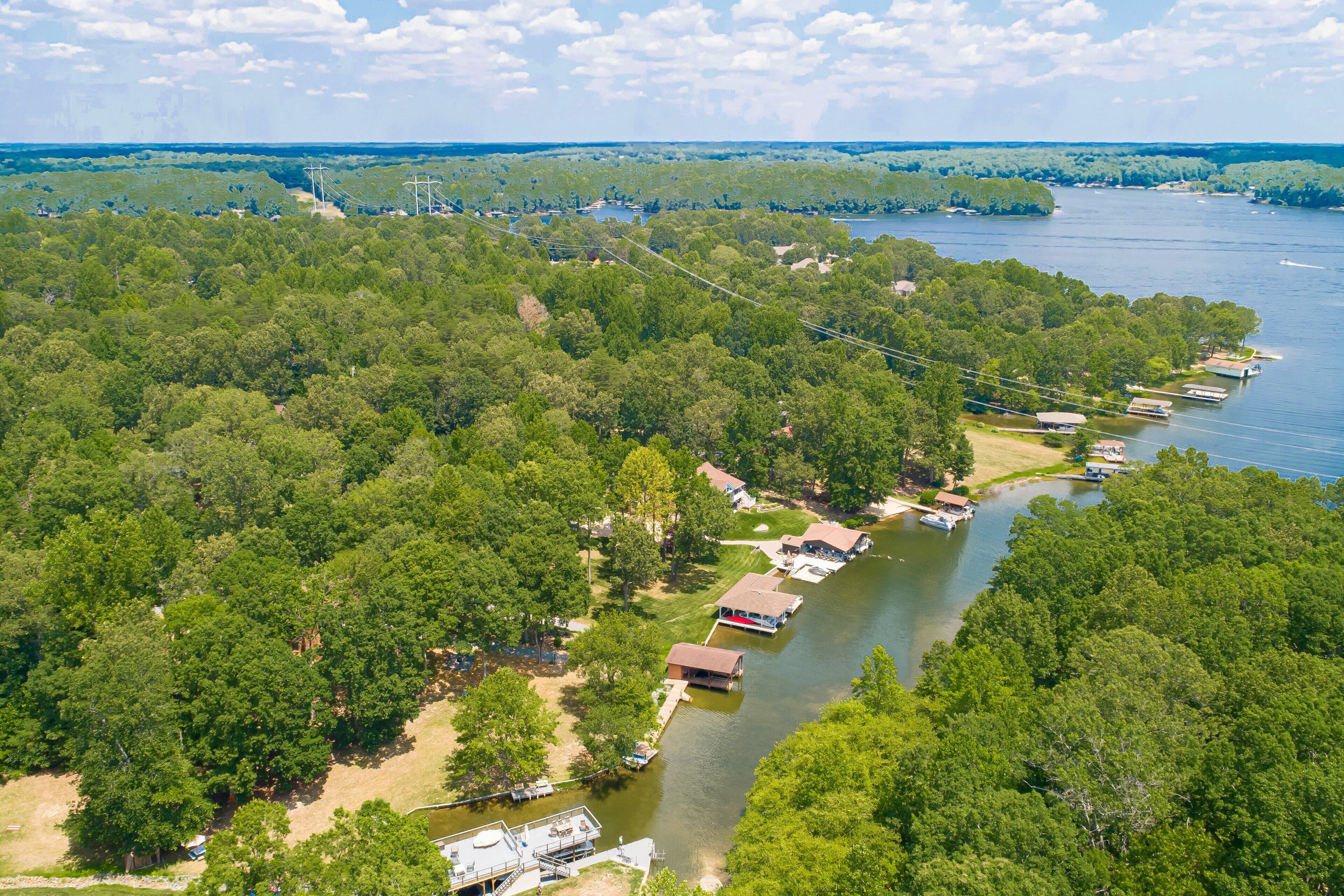 Une vue aérienne d’une propriété au bord du lac, où plusieurs bateaux sont amarrés le long de la rive, le tout étant entouré d’une forêt dense.