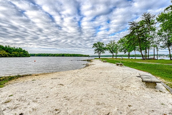 A sandy beach with a bench, a grassy area with trees, and a lake under a cloudy sky. - Lakefront Home With Kayaks, Private Patio, air Hockey, Grill, & Balcony (Mineral)