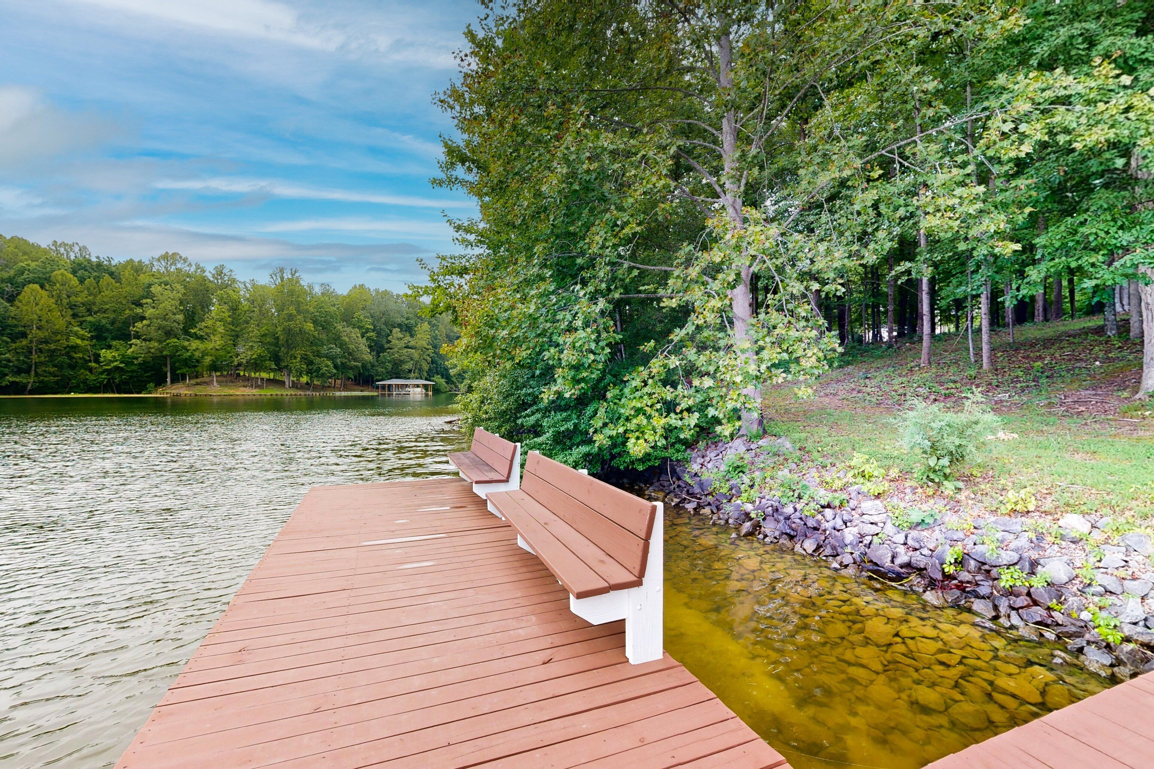 A wooden dock with benches extending over a calm lake, surrounded by dense trees and a rocky shoreline.
