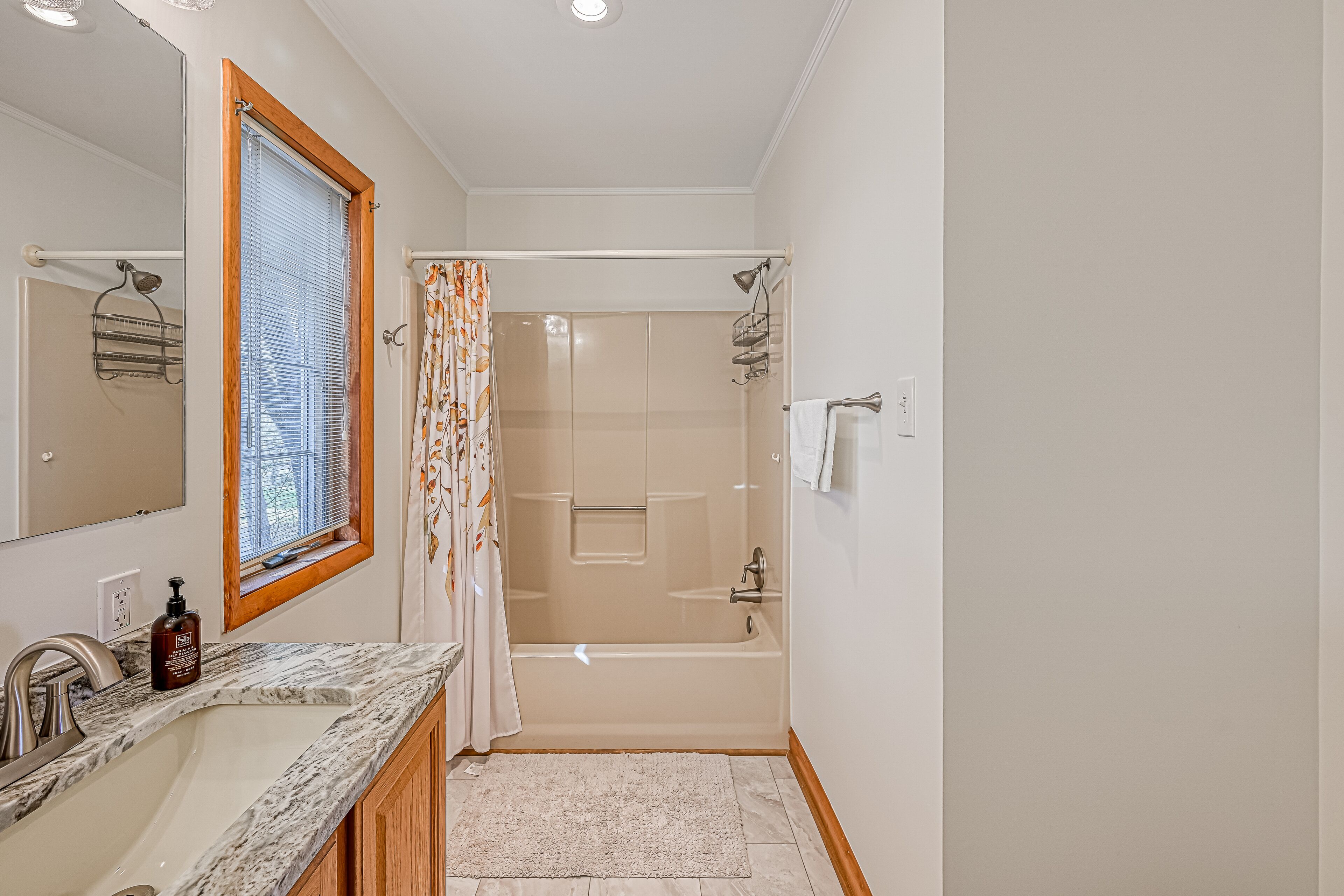 A bathroom with a sink, mirror, shower, and a window with blinds.