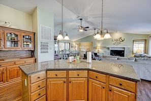 A spacious kitchen with wooden cabinets, granite countertops, and a central island. There is a dining area with a sofa and a fireplace. - Lakefront Retreat With a Large Yard, Stone Fire Pit, Gazebo, Deck, & Dock (Bumpass)