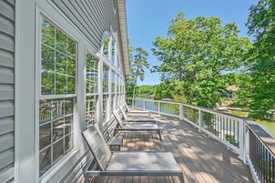 Una terraza de madera con barandillas blancas, ventanas amplias y vistas a un lago y árboles.