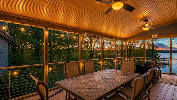 Uma área de refeições ao ar livre coberta, com mesa e cadeiras, luzes de festa e vista para a água.