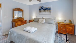 Quarto com cama, ventilador de teto, mesa de cabeceira com luminária, cômoda e um quadro na parede.