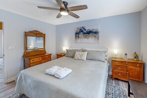 Een slaapkamer met een bed, een plafondventilator, een nachtkastje met lamp, een dressoir en een schilderij aan de muur.