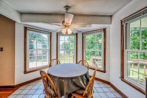 A dining area with a round table, wooden chairs, a ceiling fan, and large windows with a view of greenery. - Lakefront Home in Private Location With Dock, Firepit, Game Room & Pickleball (Mineral)