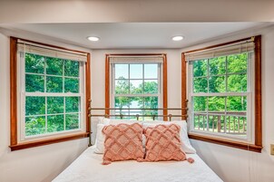 Un dormitorio con una cama grande, dos ventanas con marcos de madera y vistas al verdor exterior.
