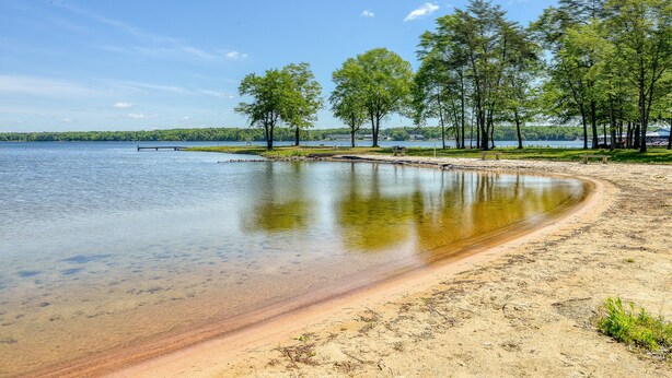 Blick auf einen See mit klarem Wasser, Sandstrand und Bäumen.