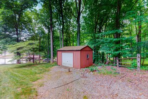 A red shed with a white garage door, surrounded by a gravel path and a wooden fence, set in a wooded area with trees and a lake in the background. - Lakefront Retreat W/private Dock, Fast Wifi, gas Grill & Central AC, Dogs (Mineral)