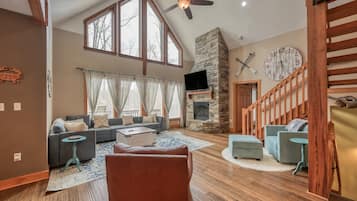 A spacious living room with a stone fireplace, large windows, a ceiling fan, and a staircase.