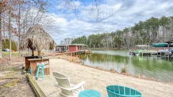 A lakeside deck with chairs, a small hut, and a dock extending into the water.