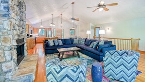 A spacious living room with a stone fireplace, a large sectional sofa, a coffee table, and a ceiling fan. - Lakefront Home With Deck, hot Tub, Game Room, Kayaks, & Fireplace (Bumpass)