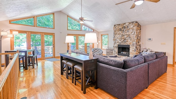 A spacious living area with a stone fireplace, wooden flooring, and large windows. - Lakefront Home With Deck, hot Tub, Game Room, Kayaks, & Fireplace (Bumpass)