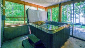 A hot tub in a covered outdoor area with large windows, wooden beams, and a view of a forest. - Lakefront Home With Deck, hot Tub, Game Room, Kayaks, & Fireplace (Bumpass)
