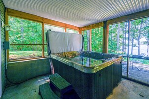 Un jacuzzi en un área exterior techada con grandes ventanales, vigas de madera y vista a un bosque.