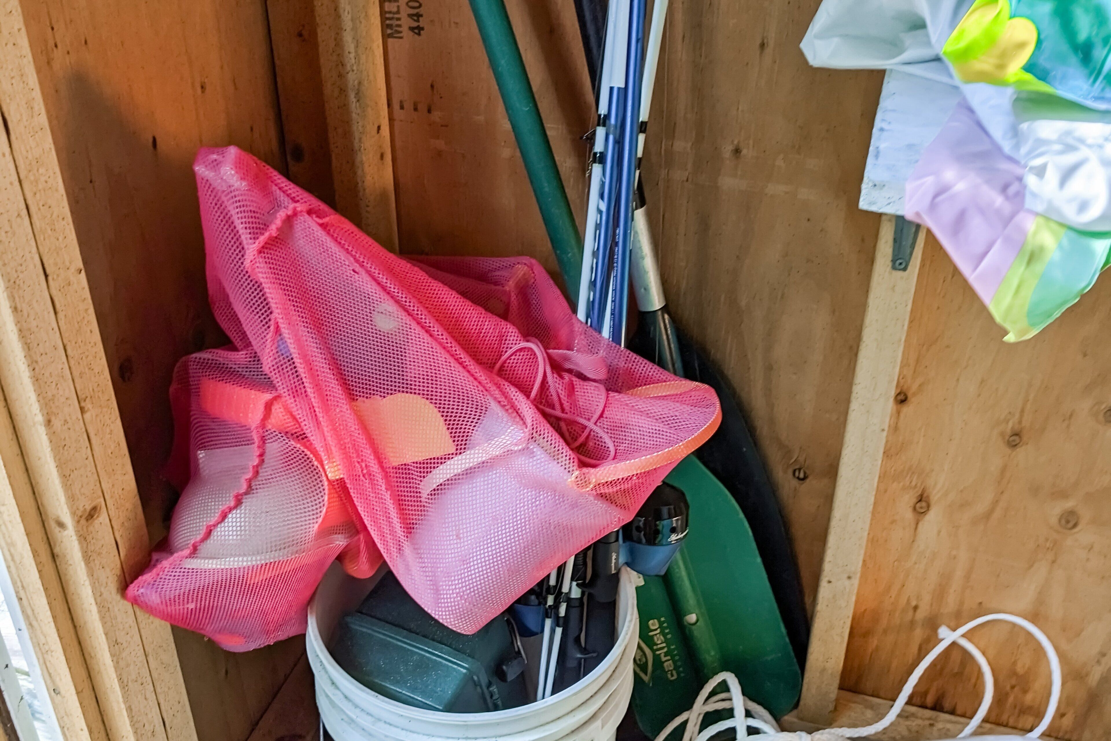 A wooden storage area with a white bucket, a green tool, and a pink mesh bag.