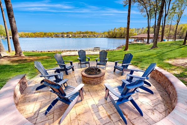 A circular outdoor seating area with fire pit, surrounded by chairs, overlooking a lake with a dock and houses in the background. - Lakefront Home With Private Dock, Kayaks, Beach, Badminton, Firepit, & Deck (Mineral)